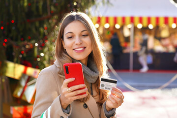 Happy smiling young woman paying her gifts online with smartphone and credit card with Christmas markets and tree with boxes on the background