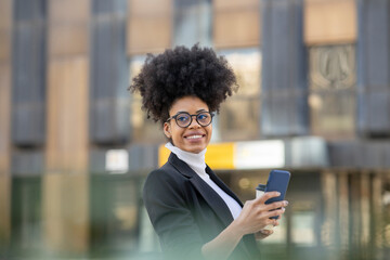 Cheerful black businesswoman with smartphone and takeaway coffee in city