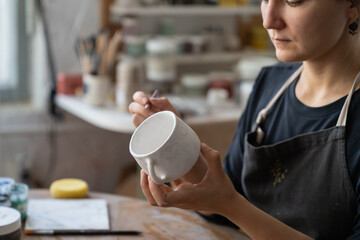 Craftswoman creates new masterpiece in work studio making sketch of future pattern on white mug....