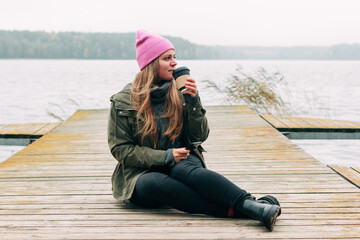 Woman in hat outdoor in the forest sitting on the bridge drinking a cup of hot coffee. Beautiful female relax and enjoy outdoor activity lifecycle in autumn nature 