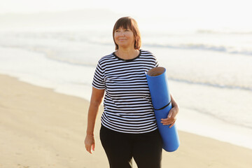 Middle age woman holding a sports mat and preparing to practice yoga outdoors on sea beach. Happy mature overweight woman exercising on seashore. copy space. Meditation, yoga and relaxation concept. - obrazy, fototapety, plakaty