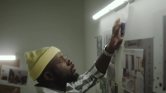 African American designer pinning printed interior plan to wall while working in creative studio