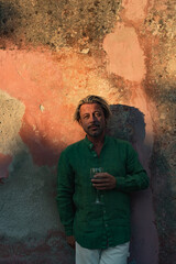 Blond tanned man in green shirt stands with glass of wine in front of weathered pink plaster wall in late afternoon sunlight.