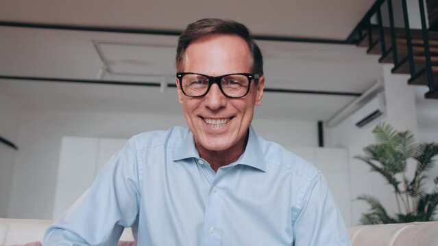 Cheerful happy mature 60s man in glasses waving hello at camera, smiling, laughing, speaking on video call in kitchen. Senior blogger, vlogger, grandfather home head shot portrait