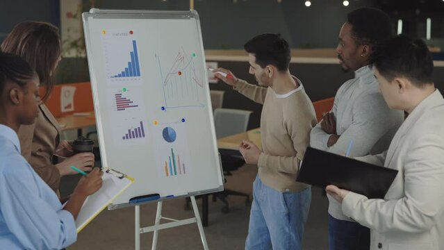 Casually dressed male coach drawing graphs on flip chart while consulting diverse staff employees in office. Concept of business, office people and teambuilding.