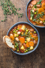 Minestrone vegetable soup on a wooden textured background with pieces of toasted bread and herbs. Simple vegan rustic Italian dish, classic of Italian cuisine, delicious healthy Mediterranean lunch