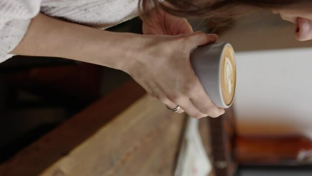 Vertical shot of attractive young woman hands hold coffee with latte art and enjoy it near the wooden table in front of window in the loft cafe 4k