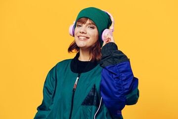 a happy, joyful woman in a green jacket and hat enjoys music standing in pink headphones on a yellow background holding them with her hands and looking at the camera