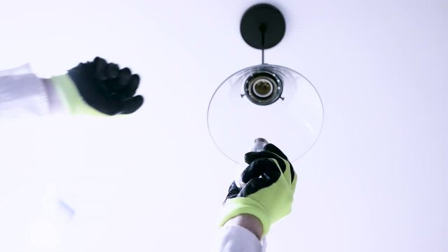 A man with one hand in gloves unscrews the lamp from the parton near the hanging ceiling lamp. Unscrewing a burnt out light bulb, replacing the bulb with an electrician in slow motion. 