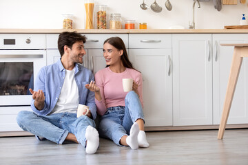 Cheerful smiling young wife and husband with cups of hot drink sitting on floor and talking at kitchen