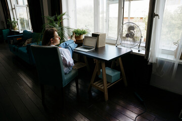 A woman is sitting at a desk in a home office and reading a book, there is a laptop on the table. 