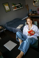 A woman is sitting at a desk in a home office and resting, holding a book in her hand