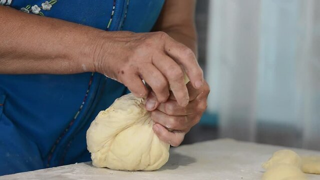 Close-up of the hands of an elderly woman in a blue dress pinching off raw dough to form buns. Culinary traditions, family recipes.