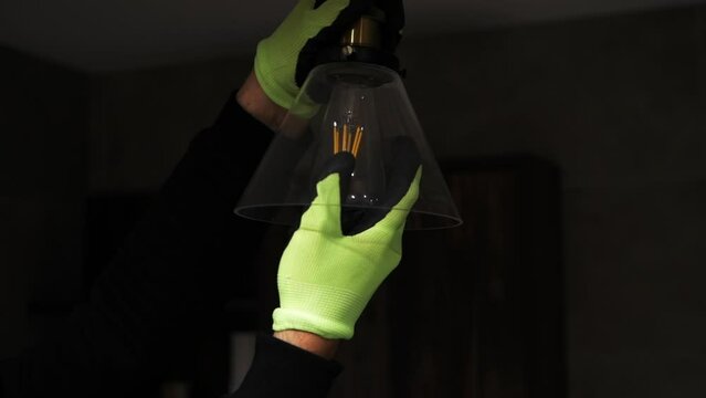  A man in gloves unscrews a lamp from a parton near a hanging ceiling lamp. Unscrewing a burnt out light bulb, replacing the bulb with an electrician in slow motion. The concept of saving electricity 