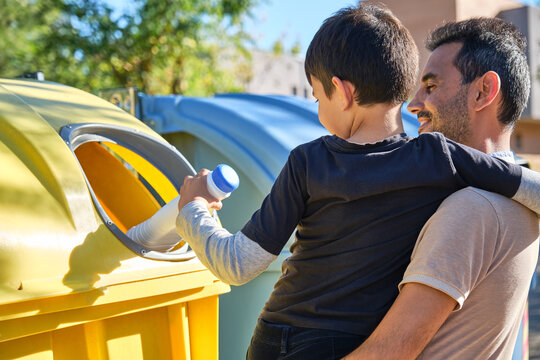 man with his young son in his arms helping to throw an empty container into the yellow recycling bin