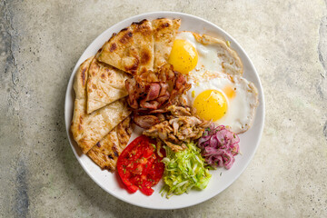 Greek Breakfast on a plate with two eggs, bacon, pita, vegetables top view on stone table