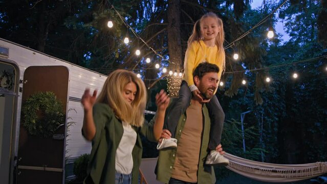 Caucasian family have party in forest travel in camping dancing with music near van. Bearded daddy man father holding kid daughter on neck shoulders dace with wife woman mother movement jumping fun