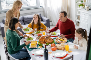 Cheerful teenager sitting near multiethnic family and tasty thanksgiving dinner at home