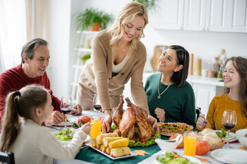 Smiling multiethnic family looking at woman with tasty turkey during thanksgiving dinner at home