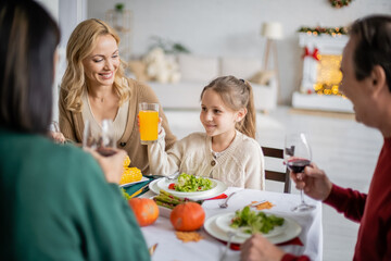 Obraz na płótnie Canvas Smiling girl holding orange juice near mother and blurred grandparents during thanksgiving celebration