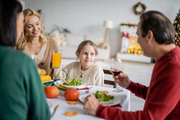 Smiling kid with orange juice looking at blurred grandparent during thanksgiving celebration at home