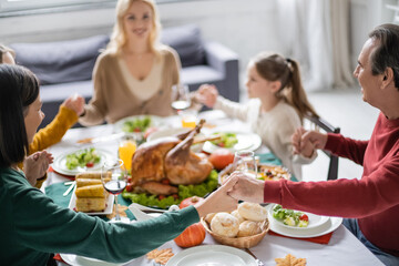 Interracial grandparents holding hands near blurred family and thanksgiving dinner at home