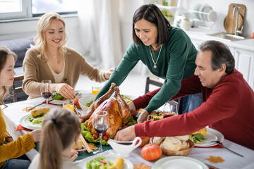 Multicultural woman putting turkey on table near thanksgiving dinner and family at home
