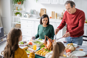 Mature multiethnic grandparent looking at delicious turkey near family celebrating thanksgiving at home