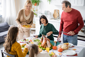 Smiling woman holding delicious pie near multicultural family and children during thanksgiving dinner