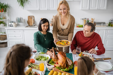 Smiling woman holding thanksgiving pie near multicultural parents and kids at home