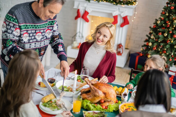 smiling senior man serving grilled corn near blurred granddaughter during christmas celebration