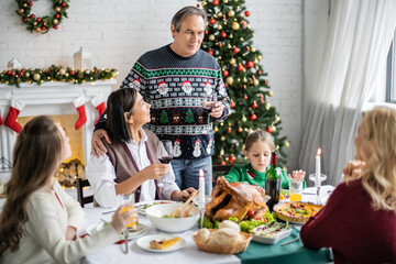 senior man toasting with glass of red wine near multiethnic family during festive christmas dinner