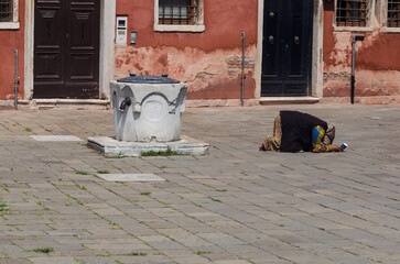 Una mendicante chiede l'elemosina inginocchiata davanti a una vera da pozzo in una piazza di Venezia