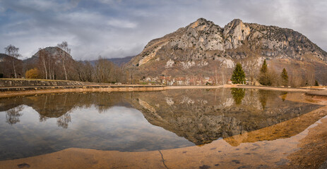 Mountain peak and trees reflecting in artificial pond. Surba town hiding behind trees at the...