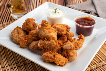 Chicken nuggets on a white square plate with two bowls of red sauce and white cheese sauce and glass of beer