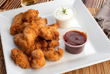 Chicken nuggets on a white square plate with two bowls of red sauce and white cheese sauce and glass of beer