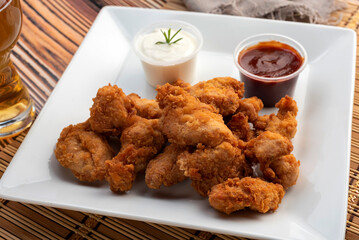 Chicken nuggets on a white square plate with two bowls of red sauce and white cheese sauce and glass of beer