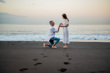 Yong man making a marriage proposal on a beach at sunset to a young beautiful woman. Standing on on...