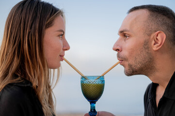 Fototapeta premium Young couple sitting on a beach at sunset and looking at each drinking from one glass with a drinking straw