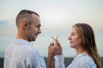 Fototapeta premium Young couple holding an Italian breadstick in their hands on a beach at sunset