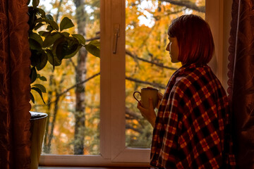 Obraz na płótnie Canvas Autumn coffee. A beautiful girl with a cup of coffee in her hands sits and looks out the window. Cozy autumn composition. Copy space.