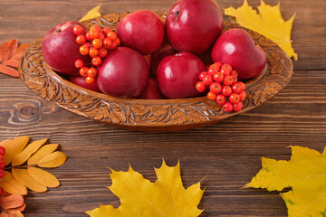 Naklejka na ściany i meble Autumn ripe red apples and mountain ash in a brown wooden bowl on a brown wooden table, next to orange autumn maple leaves. Autumn background