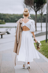 Fototapeta na wymiar A beautiful blonde bride walks through the park with a bouquet of flowers, with an autumn coat on her shoulder. Wedding photography, portrait of a beautiful model.