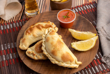 Argentinian meat or chicken empanadas on a round wooden plate with lemon wedges and bowls of hot sauce