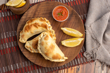 Argentinian meat or chicken empanadas on a round wooden plate with lemon wedges and bowls of hot sauce
