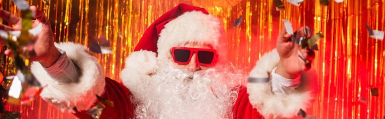 Santa claus in sunglasses and costume throwing confetti during party near tinsel, banner.