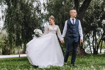 A stylish groom in a blue vest and a beautiful smiling bride are walking in the park on the green grass, holding hands. Wedding photo of newlyweds in love.