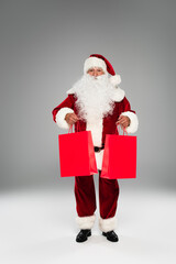 Full length of santa claus holding red shopping bags on grey background.