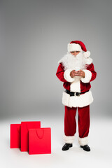 Full length of father christmas using cellphone near shopping bags on grey background.