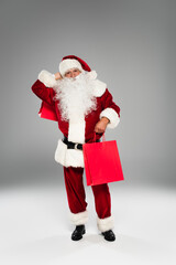 Full length of santa claus in costume holding purchases on grey background.